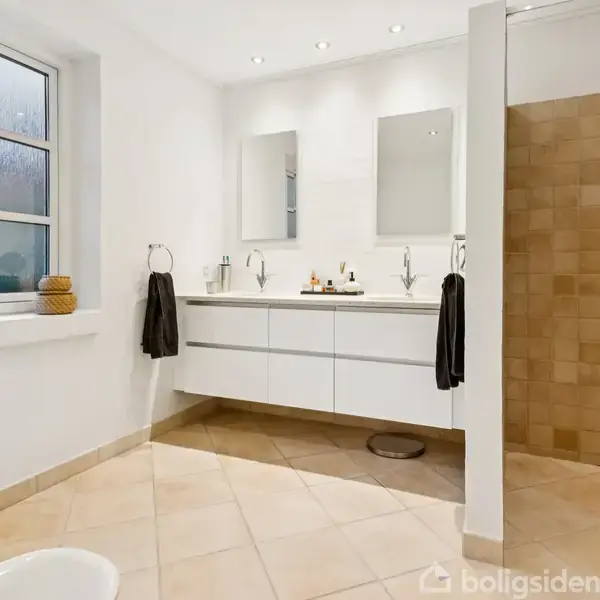 Badeværelse med dobbel håndvask på en vægmonteret skab, to spejle ovenover. Til højre en bruser med beige fliser. Vindue til venstre, planter og kurv på vindueskarmen.