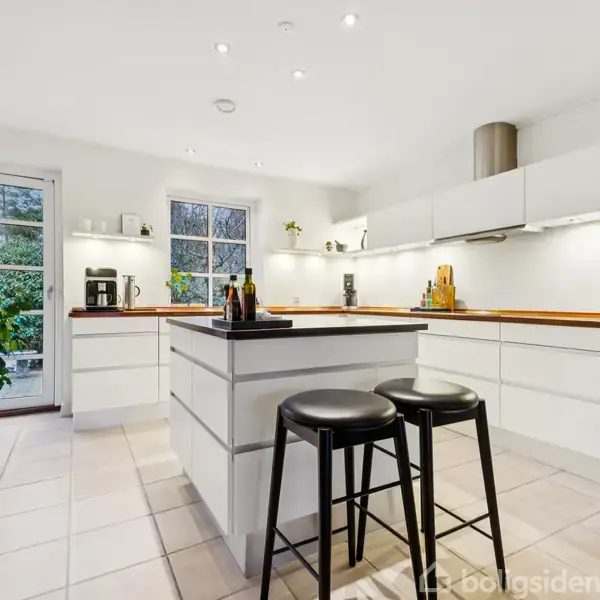 Stue med flisegulv og lyse vægge. Et stort spisebord med stole står midt i rummet. En grøn plante står ved væggen nær et maleri.