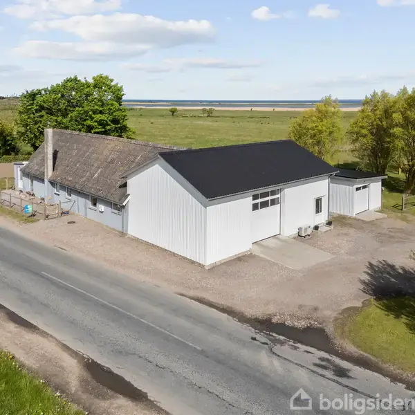 Hvid bygning langs vej, flankeret af træer og marker. Bygningens tag er sort, og der er en mindre udhusbygning ved siden af. Vejforløb strækker sig gennem grønne omgivelser.