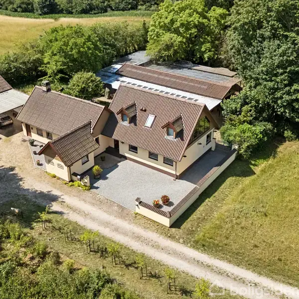To huse med røde tage og hvide vægge står ved en grusvej. Omgivet af træer og marker skaber de en idyllisk landlig atmosfære.