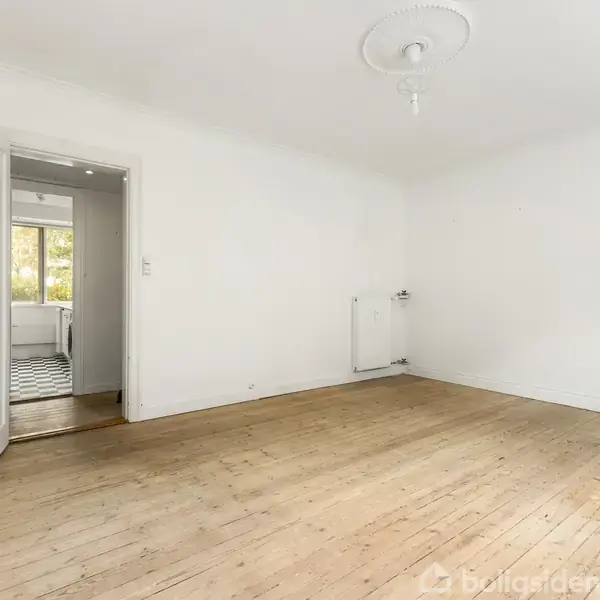 Et tomt rum med trægulv og hvidmalede vægge. En åben dør fører til et tilstødende rum med flisegulv. Et radiator er monteret på væggen. Ingen tekst.