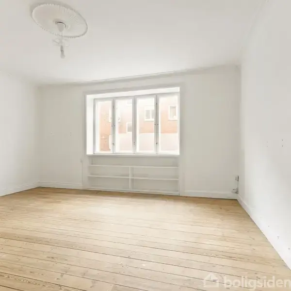 Et tomt værelse med et trægulv har en stor vinduesparti, der lukker lys ind. Væggene er hvide, og der er en enkelt lampeudtag i loftet.