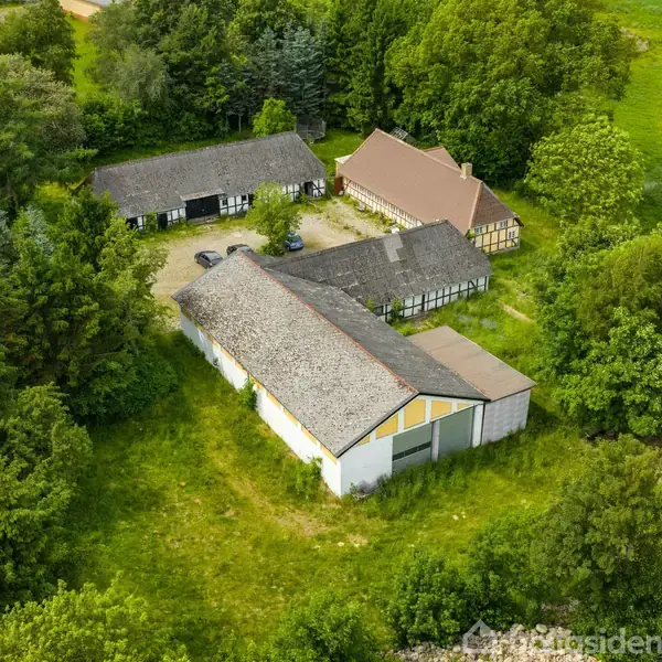 Luftfoto af gård med flere bygninger omgivet af tæt skov og grønne marker. Bygningerne er placeret tæt sammen, og der ses en gårdsplads mellem dem.