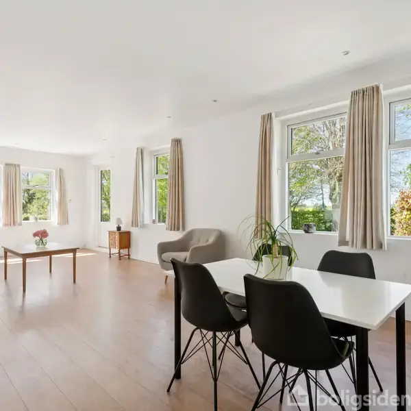 Spisestue med bord og fire stole står ved siden af et stort vindue med udsigt til grønne træer. Sofa og sofabord er placeret modsat, og rummet er lyst.