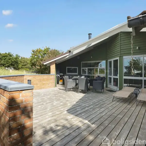 Terrasse med trædæk og loungemøbler foran et moderne hus med grønne vægge. Omgivende natur med græs og træer. Klar blå himmel giver indtryk af solrig dag.