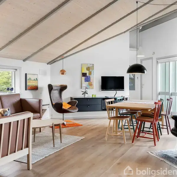 Stue med moderne møblering: en brun sofa og lænestol, et spisebord med farverige stole. Væggene prydes af kunst, og et TV hænger på den hvide væg. Store vinduer slipper lys ind.