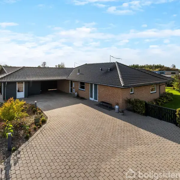 En moderne, murstensvinkelhus står på en stenbelagt indkørsel, omkranset af velplejet hække og have i en rolig forstad.