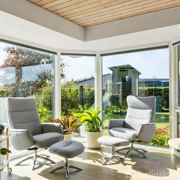 To moderne lænestole står i et lyst udestuemiljø med store vinduer. Udenfor ses en have med grønne planter og et lille skur. Rummet har træloft og flere potteplanter.