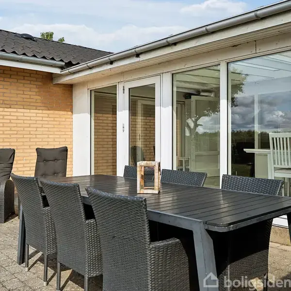 Udendørs terrasse med bord og otte stole i sort flet. Til venstre står to liggestole. Terrassen grænser op til en gul murstensbygning med store vinduespartier.