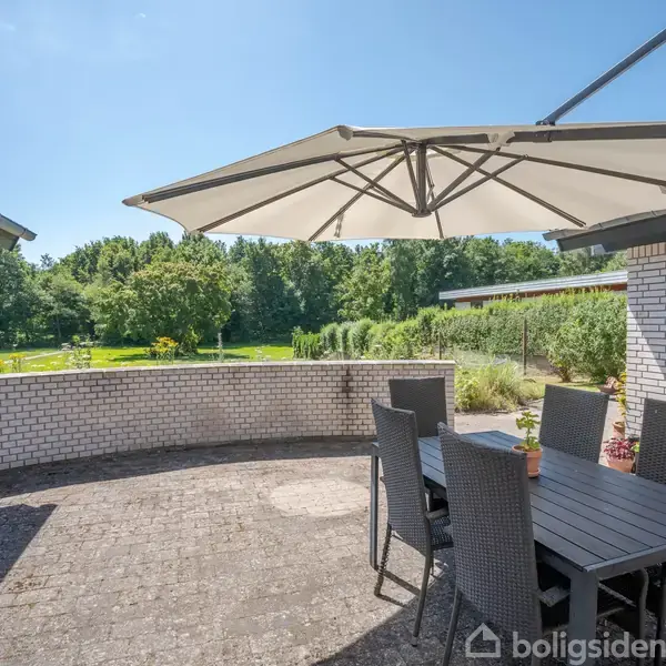 Spisebord med seks stole står under en stor parasol på en fliseterrasse ved et hvidt murstenshus. Græsplæne og træer ses i baggrunden under en blå himmel.