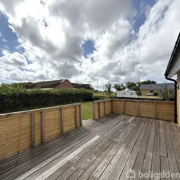 Terrasse med træbrædder omkranset af et lavt hegn; baggrunden viser en grøn have, en hæk, og huse under en delvist skyet himmel.
