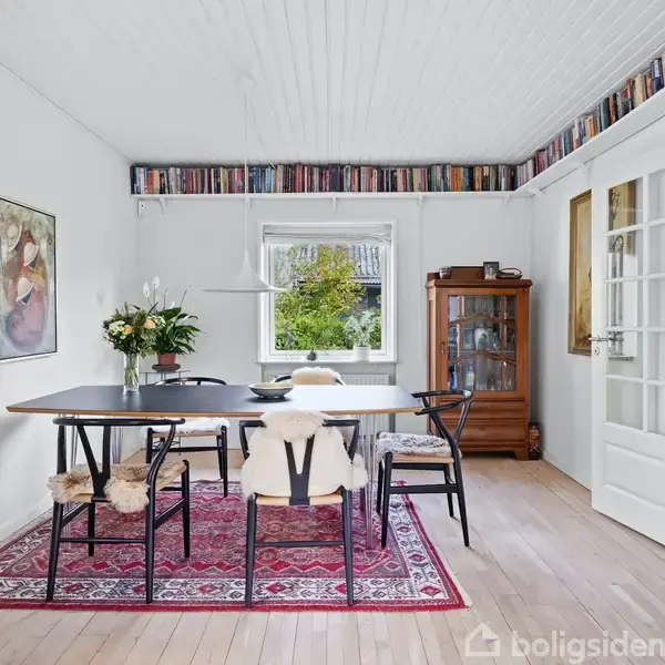 En spisestue med et stort bord omgivet af stole med pelsbetræk. En vase med blomster står på bordet. Rummet har trægulv, en rød orientalsk tæppe, og en bogreol langs loftet.