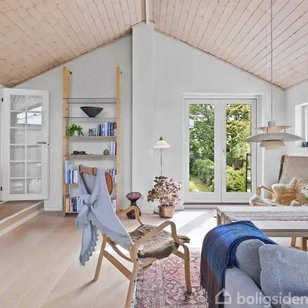 Stue møbleret med moderne stole og sofaer. Sollys strømmer gennem store vinduer. Reoler indeholder bøger og dekorationer. Et tæppe dækker gulvet, og grønne planter tilfører liv.