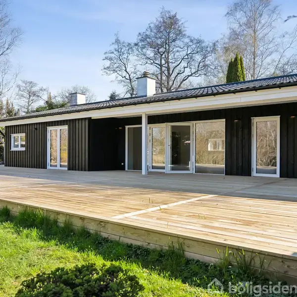 Sort træhus med store vinduer står stille. Huset har en rummelig træterrasse foran og er omgivet af en grøn have med træer i baggrunden.