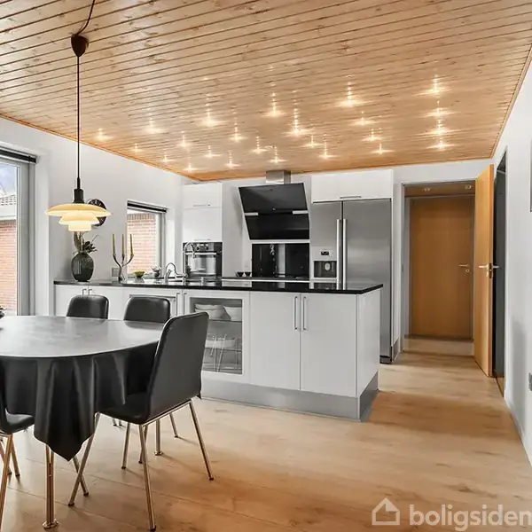 En moderne køkken- og spisestue med hvidt interiør, et rundt bord med sort dug og seks stole. Over bordet hænger en lampe. Trægulv og træloft skaber en varm atmosfære.