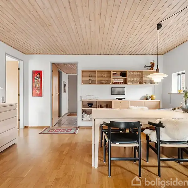 Et spisebord med sorte stole står i en lys spisestue med trægulve og matchende træloft. Malerier hænger på væggene. Skabe og hylder er langs væggene.