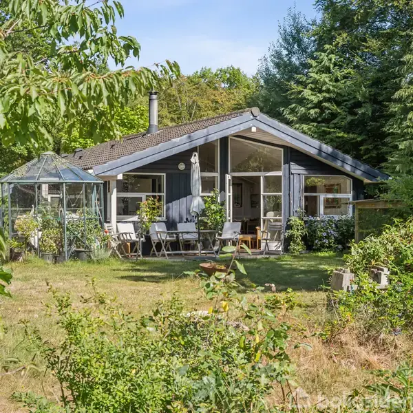 Et mørkeblåt sommerhus med store vinduer og terrasse ligger omgivet af frodig grønt og træer. En glaspavillon står til venstre i haven.