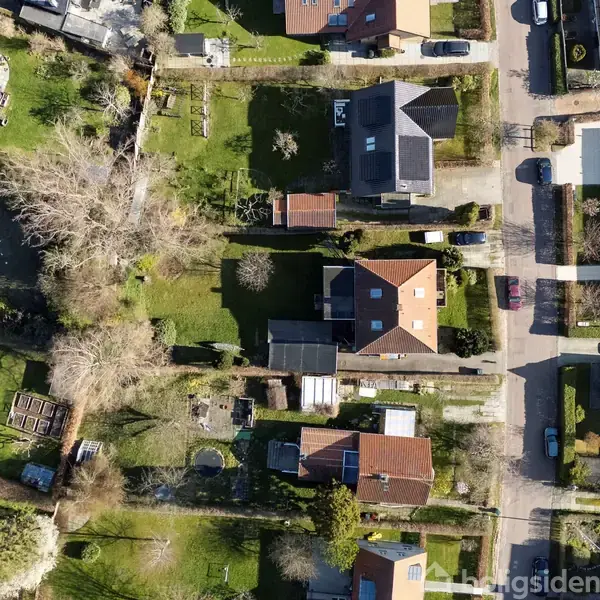 Huse set ovenfra, ligger i en forstad med grønne haver og træer. Små veje snor sig mellem bygningerne. Haverne indeholder træer og buske, og der er få parkerede biler.