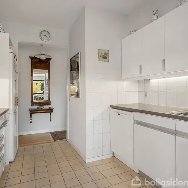 Køkken med hvide skabe og stålbordplade. Komfur og emhætte til venstre. Gang med spejl og træbænk i baggrunden. Lys flisegulv og vægmonteret ur.