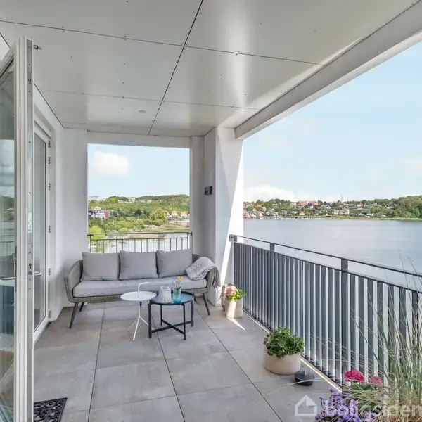 Balkon med sofa og bord står stille. Kontekst er en panoramaudsigt over sø med grønne bakker og blå himmel. Åben dør til indendørs rum.
