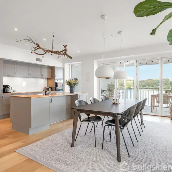 Et moderne køkken-alrum med et mørkt spisebord og seks stole, placeret på et lyst tæppe. En stor plante og åbne terrassedøre giver udsigt til en balkon og natur udenfor.