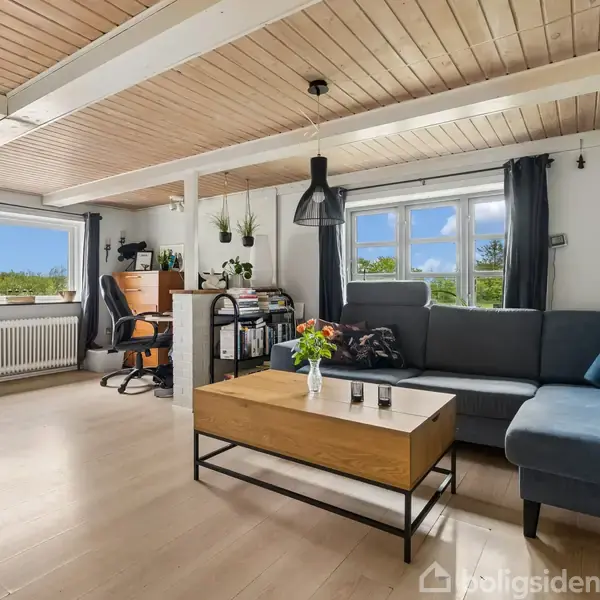 En stue har en grå hjørnesofa ved et vindue med sorte gardiner. Et træbord står foran sofaen. Til venstre er et skrivebord med en kontorstol, omgivet af lyse trægulve og hvide vægge.