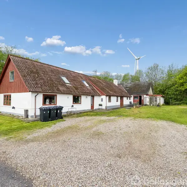 Hus med rød tag og hvid facade står på grusbelagt gårdsplads omgivet af grønt område. En vindmølle ses i baggrunden, og skraldespande står foran huset.