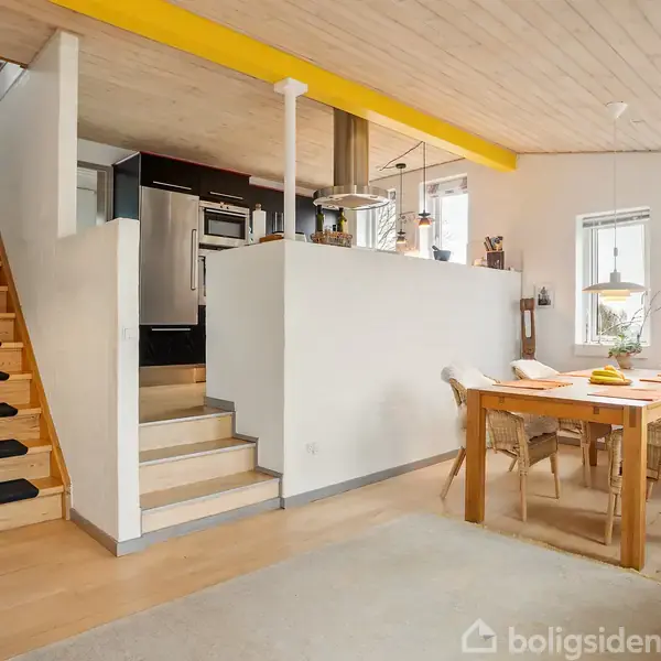 Trætrappe fører til øverste etage; spisebord med stole står i lyse, moderne spisestue med træloft, store vinduer, lysindfald og åbent køkkenområde.