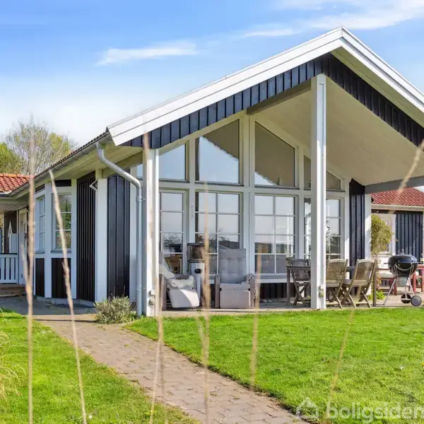 Et sommerhus i træ med store vinduer og en terrasse med havemøbler og grill. Huset står på en grøn græsplæne omgivet af buske og træer under en blå himmel.