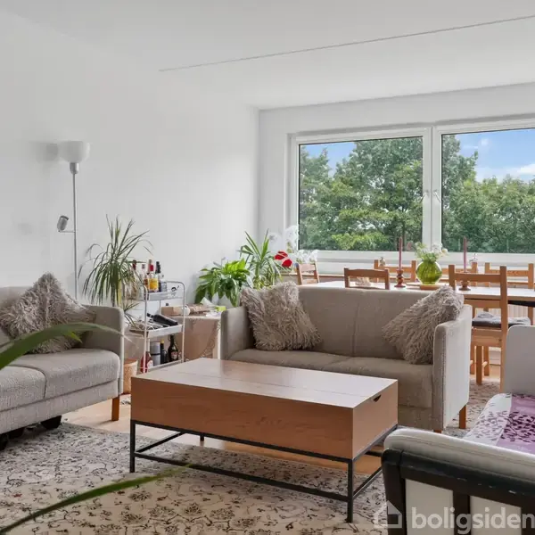 En lys stue med to sofaer og et bord i centrum. Et spisebord med stole står ved vinduet med udsigt til grønne træer. Indretningen inkluderer planter og lamper.