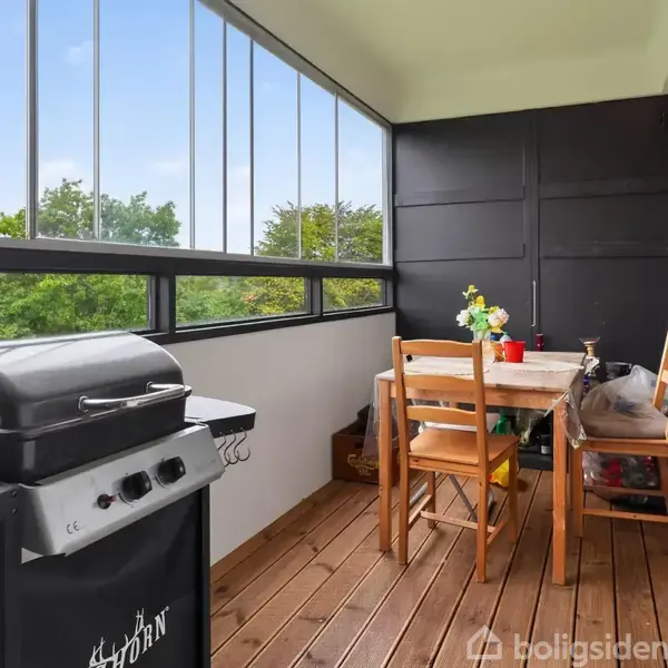 Grill står ubenyttet på altan med træplanker, omgivet af stole og et bord med blomsterarrangement. Store vinduer til højre giver udsigt over grønne træer i baggrunden.