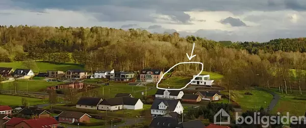 En pil peger mod et tomt byggegrund markeret med en cirkel i en boligområde med huse og en skovbaggrund.