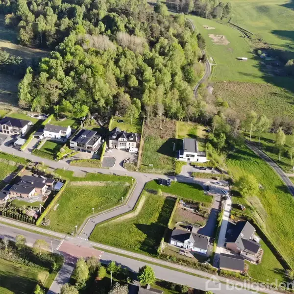 En række huse er omgivet af grønne marker og skov. Veje forbinder husene, som ligger langs en bred grøn skovkant. Området er præget af en landlig atmosfære.
