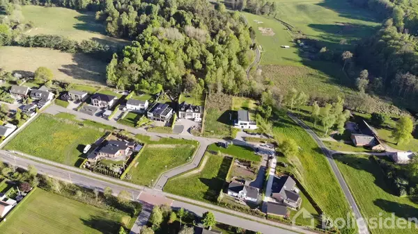 Flyfoto viser række af moderne huse langs en vej i et grønt, skovrigt område omgivet af marker og stier.