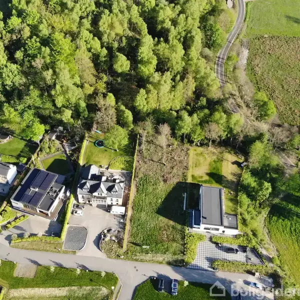 Huse set oppefra omgivet af grønne områder og en skov. En vej løber gennem området, mens stier og åbne marker ses i nærheden. Ingen tekst er fundet i billedet.