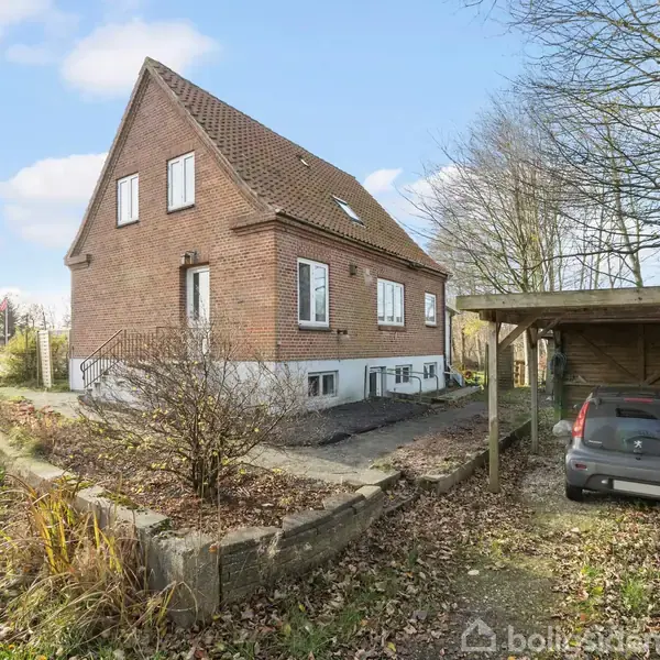 Et murstenshus med brun facade står stille. En bil er parkeret under en carport. Træer omgiver området, og en vej kan ses i nærheden til venstre.