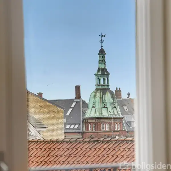 Ud af et åbent vindue ses en detaljeret kirketårn med grøn kuppel og vejrfløj, omgivet af bygningstage under en klar blå himmel.