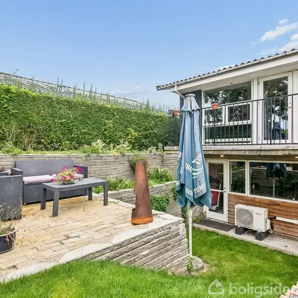 Terrasse med grå sofa og bord placeret på hævet stenterrasse i haven. I nærheden står en paraply og en dekorativ metalovn. Huset har vinduer og en terrasse med rækværk.