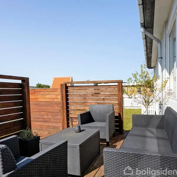 Terrassemøbler i polyrattan står arrangeret på en træterrasse. Ved siden af et hus med store vinduer er der udsigt til haveomgivelser, afskærmet af en træplankehegn under en klar blå himmel.