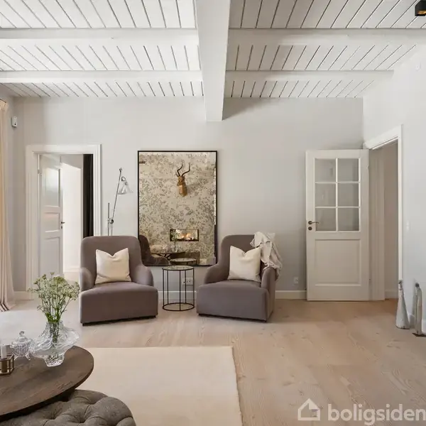 To bløde lænestole står stille ved siden af et lille bord med en lampe. De er placeret foran en dør og hvid bogreol i en lys stue.