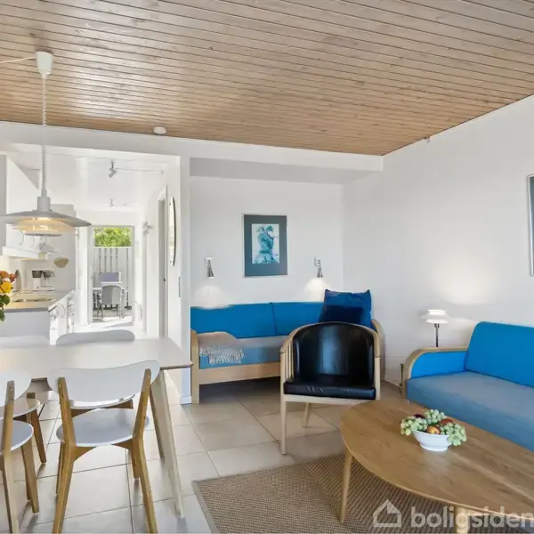 Stue med blå sofaer og et rundt sofabord. Spisebord med hvide stole står til venstre. Loftslamper hænger fra træloftet. Et åbent køkken er synligt i baggrunden.