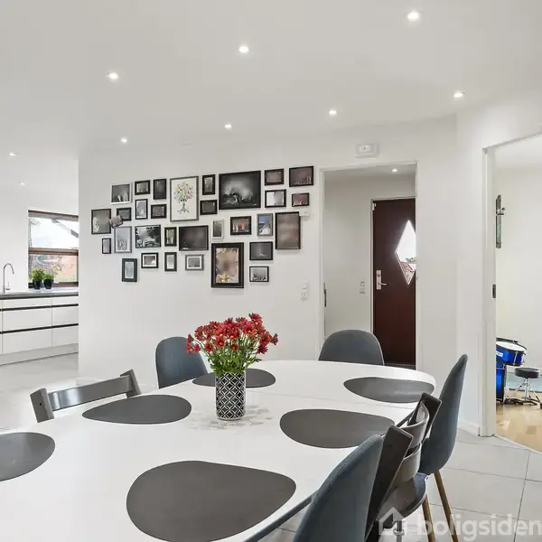 Rundt spisebord er dækket med sorte underlag og blomster i vase, i lyst spisestue. Væggen har en billedvæg. Åben adgang til køkken og gang.