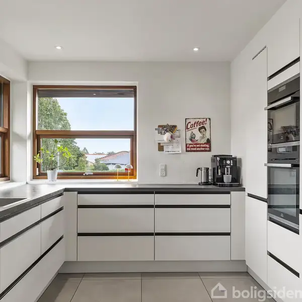 Et moderne, lyst køkken med hvide skabe og sorte greb. Kaffemaskine og plakater er på væggen. Et stort vindue giver udsigt til træer og en bygning udenfor.