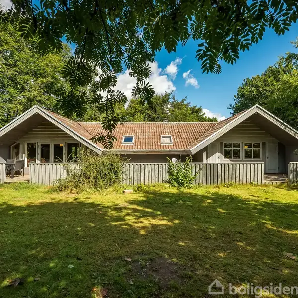 Grå træhus står stille omgivet af grønne træer og græsplæne. Sollys skinner på taget, og en terrasse omkranser huset. Klar blå himmel ses i baggrunden.