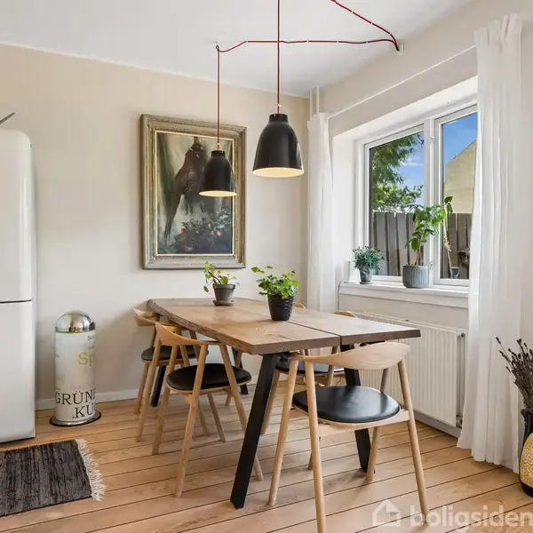 Et træbord står i en lys spisestue med fire stole omkring. En hvid SMEG køleskab er til venstre, og en plante samt et billede hænger på væggen. En stor vindue bagved.