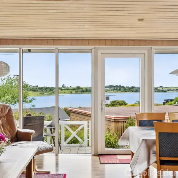 En stue med lænestol og spisebord med stole. Store vinduer og en dør åbner udsigten mod en sø og grønne bakker udenfor. Blomster på bordet.