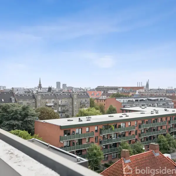En udsigt fra en altan viser et bylandskab med flade tage og flere etageejendomme. I baggrunden ses kirketårne og skorstene under en klar, blå himmel.