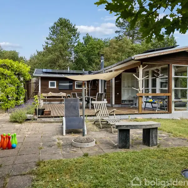 Sommerhus med store vinduer står på en flisebelagt terrasse. Havemøbler og farverige kegler til bowling er placeret udenfor i en frodig have omkranset af træer.