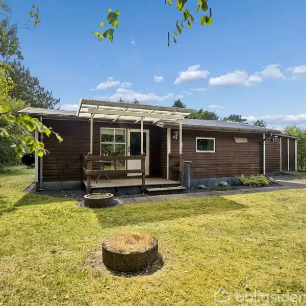 Et træhus står frit, omgivet af en græsplæne med spredt vegetation. Det har en overdækket terrasse med bord. I baggrunden ses en hæk og blå himmel med skyer.