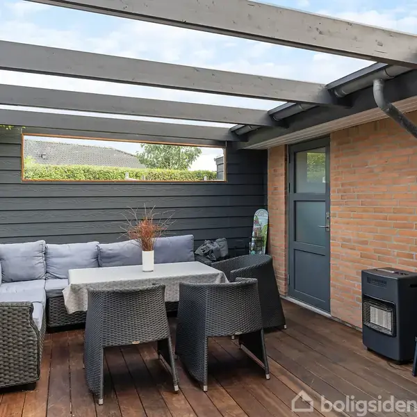 Overdækket terrasse med sofa og bord omgivet af stole. Hylder med planter og flasker står til højre. Terrassebrædder dækker gulvet, og en murstensvæg med dør ses bagved.
